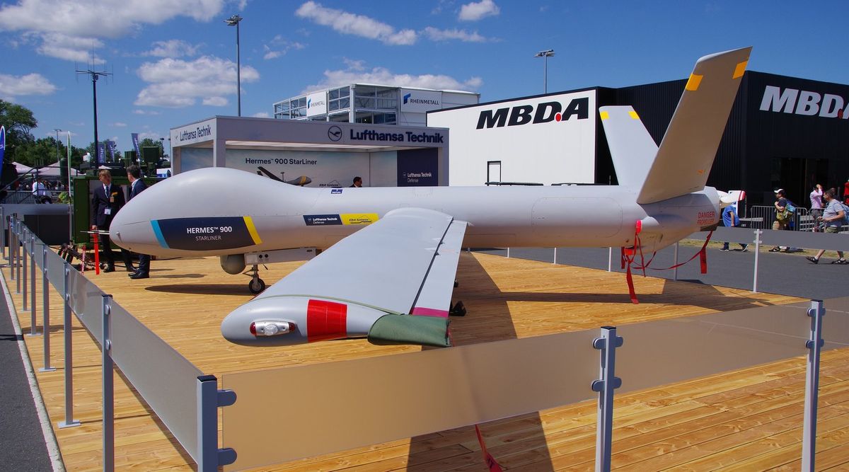 Graue Drohne mit langen Tragflächen, V-Leitwerk und der Aufschrift Hermes 900 Starliner auf einer Holzplattform hinter einer Absperrung, dahinter Messestände von Lufthansa Technik, Rheinmetall und MBDA unter blauem Himmel
