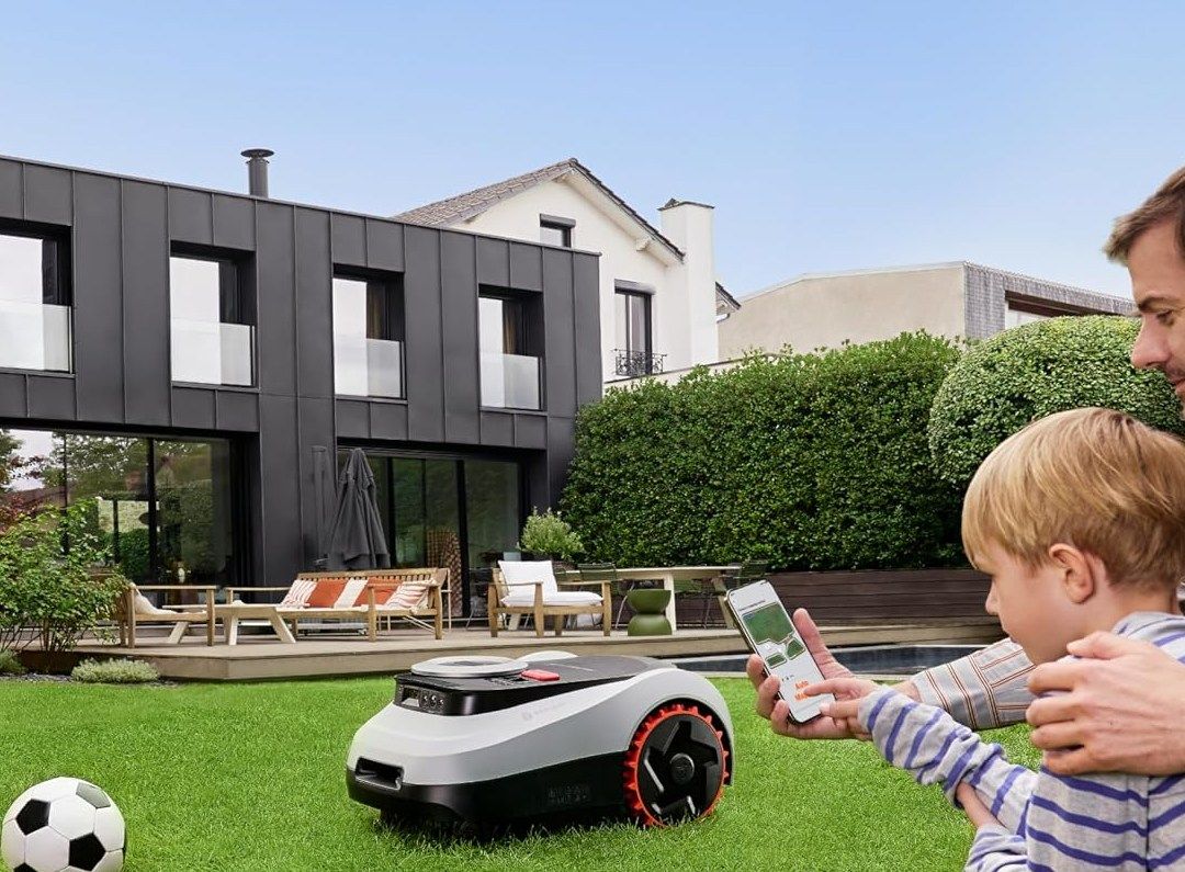 Ein Navimow-i2-LiDAR-Mähroboter fährt über den Rasen vor einem modernen Haus, rechts steuern ein Vater und sein Sohn ihn per Smartphone, links liegt ein Fußball.