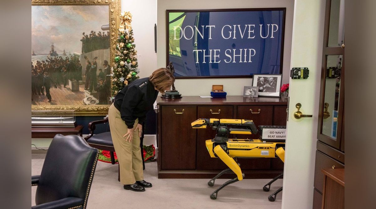 Gelber vierbeiniger Roboter Spot mit dem Schriftzug Boston Dynamics und nach vorn gestrecktem Greifarm vor einer dunklen Holzanrichte; links beugt sich eine Frau in dunkler Uniformjacke zu ihm hinunter, darüber ein Bild mit dem Schriftzug „Dont give up the ship“, links ein großes Gemälde mit Matrosen und ein geschmückter Weihnachtsbaum