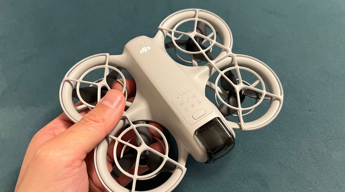 Eine Hand hält eine hellgraue DJI-Neo-Drohne mit geschlossenem Propellerschutz vor petrolfarbenem Stoff
