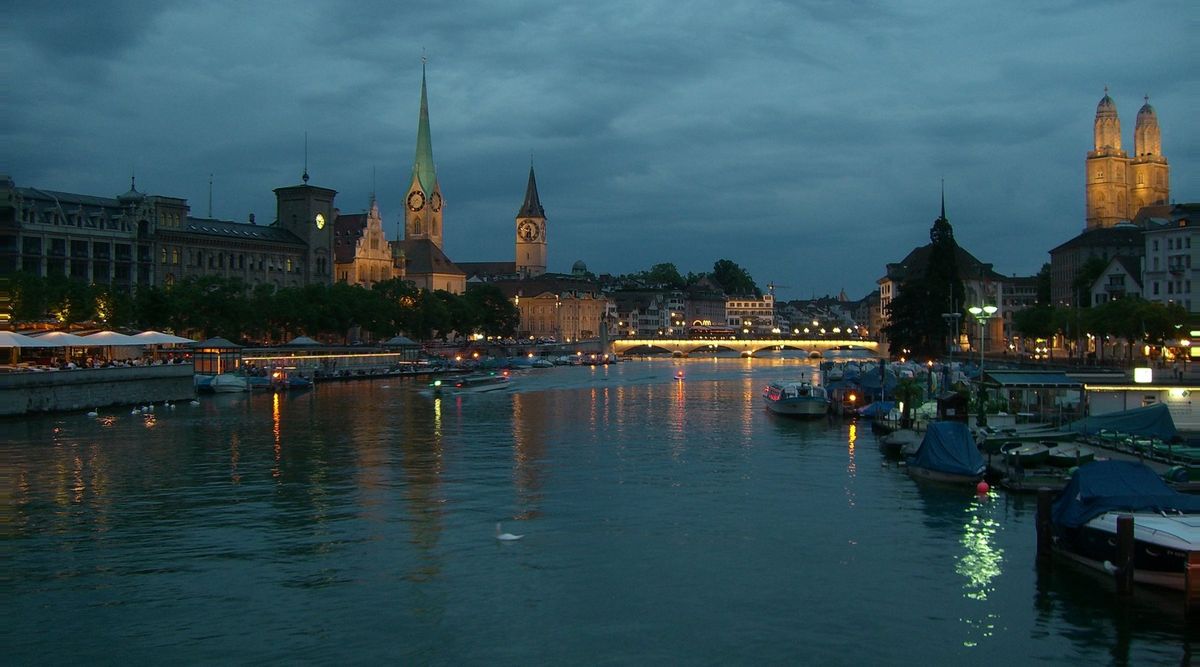 Blick über die Limmat in Zürich bei Abenddämmerung: links das Fraumünster mit grüner Turmspitze und die Kirche St. Peter mit beleuchteter Uhr, in der Mitte eine beleuchtete Brücke, rechts die beiden Türme des Grossmünsters; auf dem Wasser Boote und ein Schwan