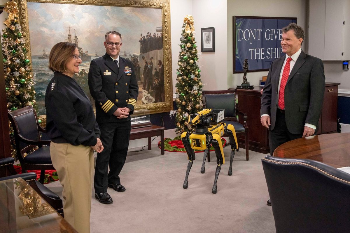 Boston Dynamics Spot mit Greifarm steht in einem Büro mit Weihnachtsbaum und Marinegemälde, links eine Admiralin und ein Marineoffizier, rechts ein Mann im Anzug.