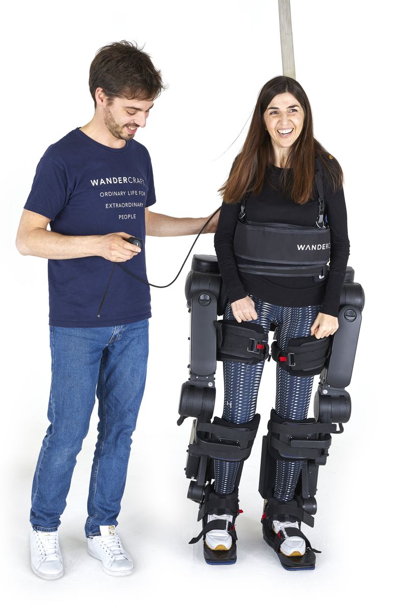 Eine Frau steht im Klinik-Exoskelett Atalante X, daneben bedient ein Wandercraft-Mitarbeiter im Firmen-T-Shirt die kabelgebundene Fernsteuerung