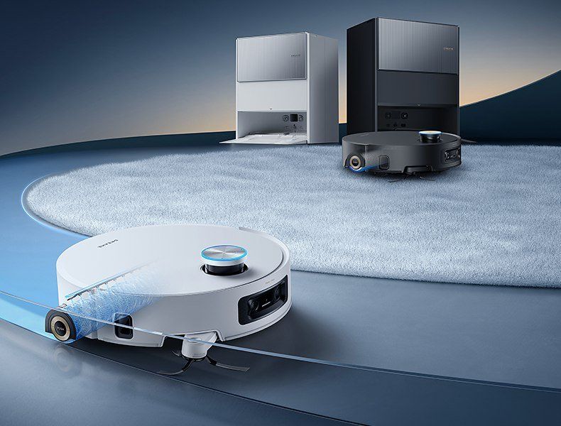 Ein weißer runder Saugroboter mit blau leuchtendem Sensorturm fährt mit einer blau angeleuchteten Walze an der Seite über einen glatten Boden, dahinter auf einem hellblauen Teppich ein schwarzer Roboter vor einer schwarzen und einer silbernen Station