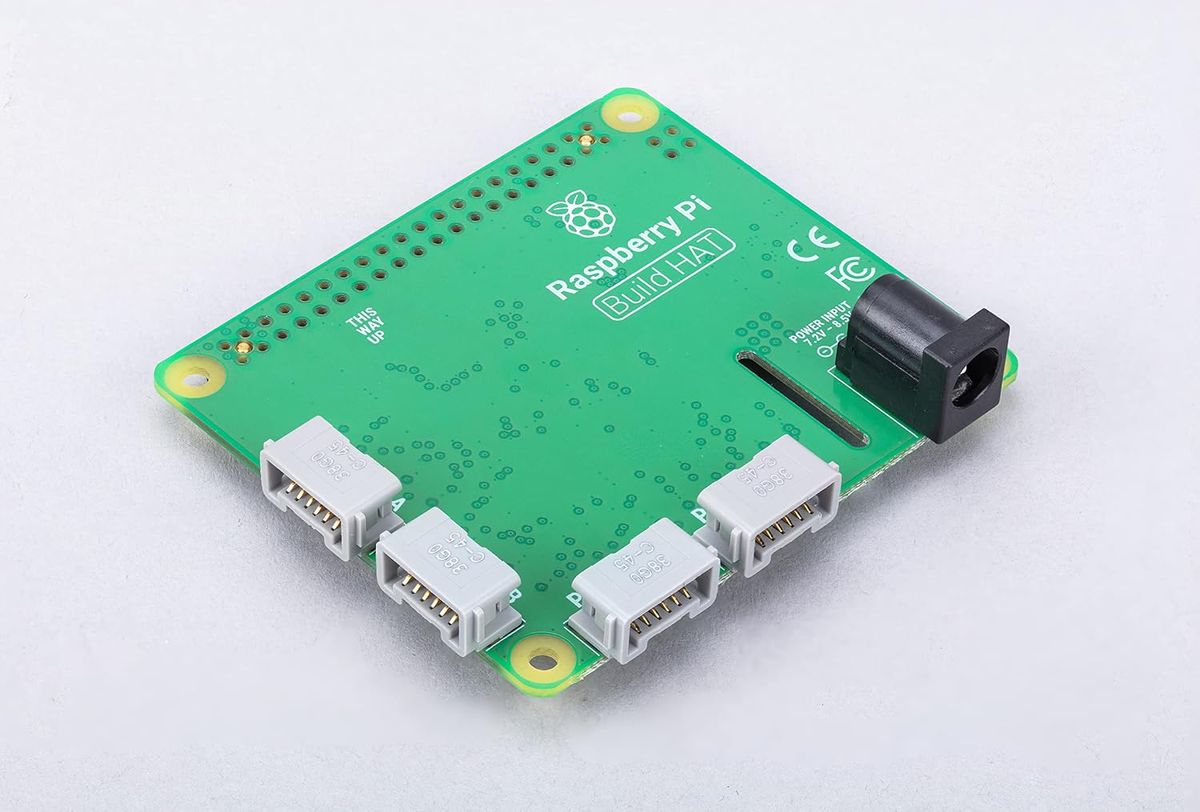 Grüne Raspberry-Pi-Build-HAT-Platine mit vier grauen LEGO-Anschlüssen und schwarzer Hohlbuchse für die Stromversorgung.