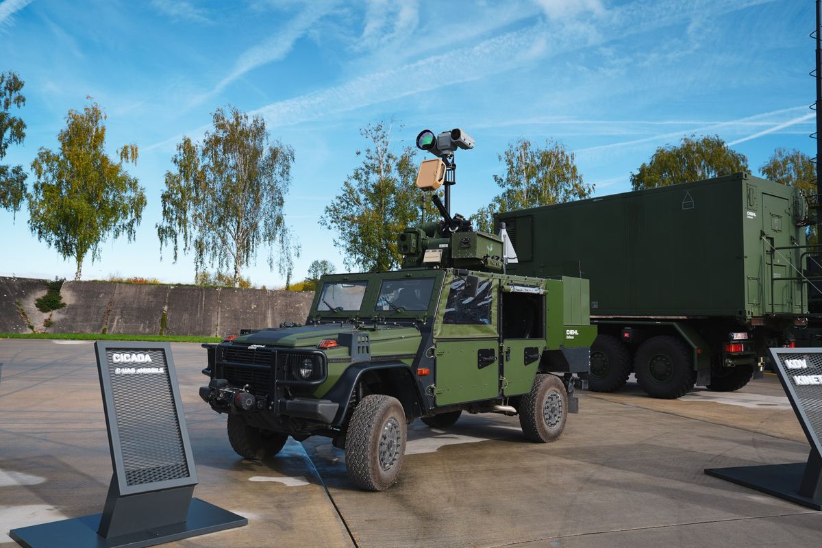 Olivgrüner Geländewagen auf Mercedes-G-Klasse-Basis mit Sensormast, auf dem ein sandfarbener Radarkasten und ein Kameramodul sitzen, und einer Waffenstation auf dem Dach; links ein Aufsteller mit der Aufschrift CICADA, rechts angeschnitten ein Aufsteller mit KDV, dahinter ein grüner Lkw mit Container, Birken und blauer Himmel mit Kondensstreifen