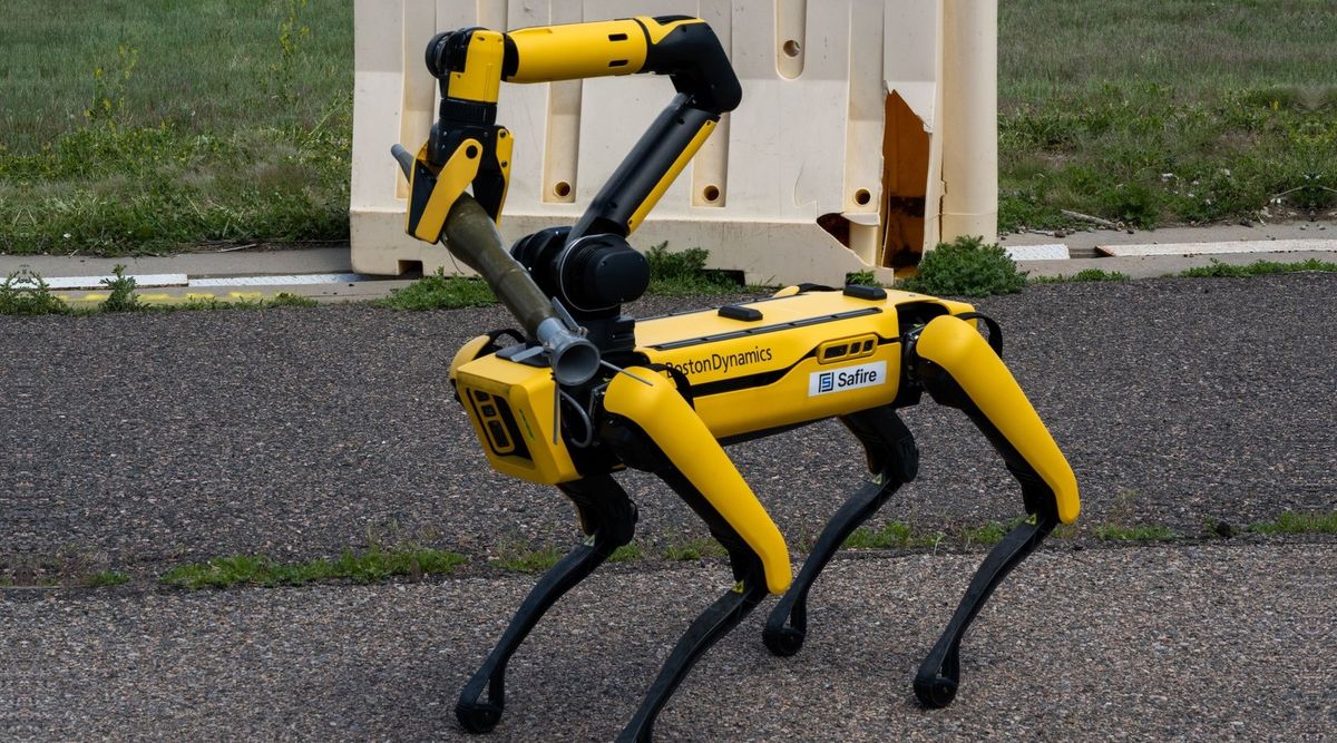 Ein gelb-schwarzer vierbeiniger Roboter mit Boston-Dynamics-Schriftzug und Safire-Aufkleber steht auf Asphalt, sein gelber Greifarm hält ein olivgrünes Rohr, dahinter eine beschädigte weiße Kunststoffbarriere und Wiese