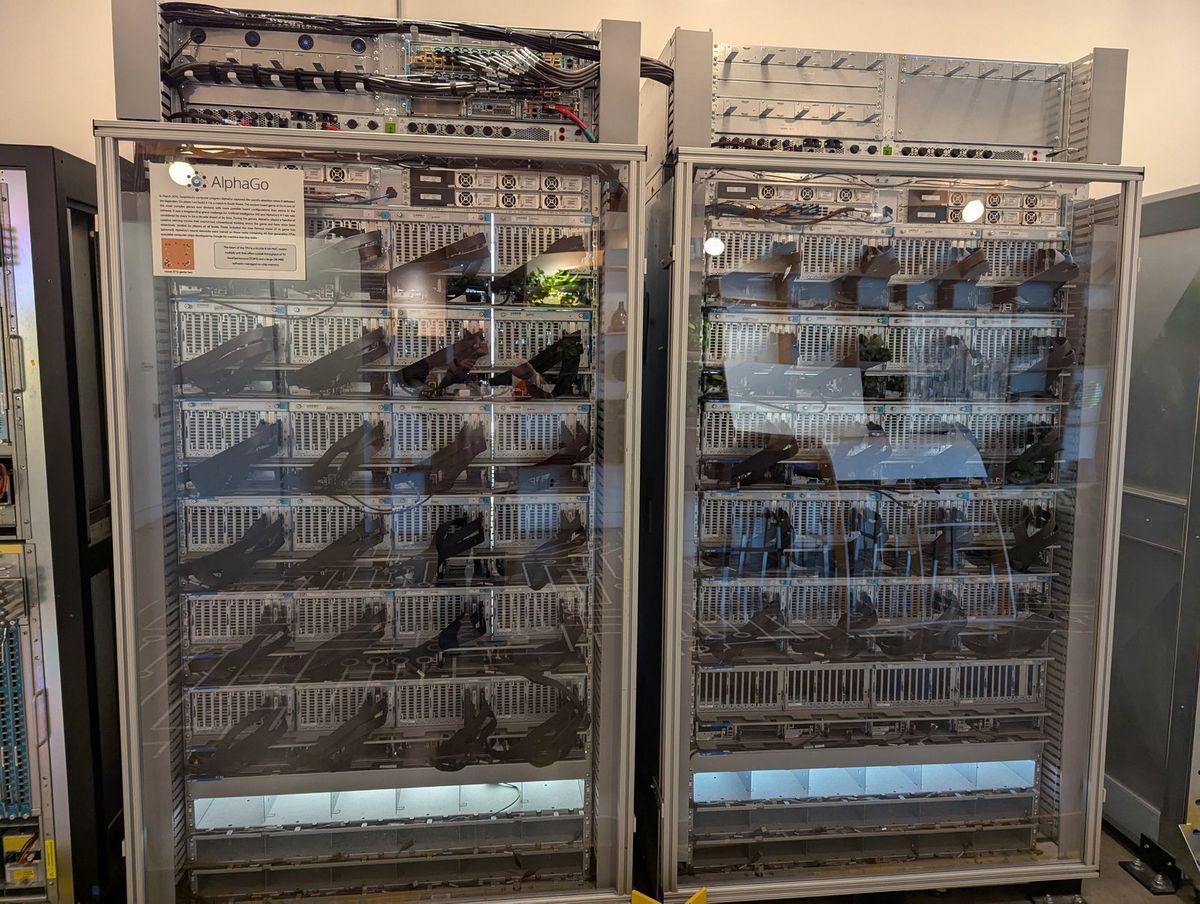 Zwei ausgemusterte Serverschränke hinter Glas, dicht bestückt mit Rechenmodulen und Kabelbündeln, links ein Schild mit dem Schriftzug AlphaGo und einer Erklärtafel