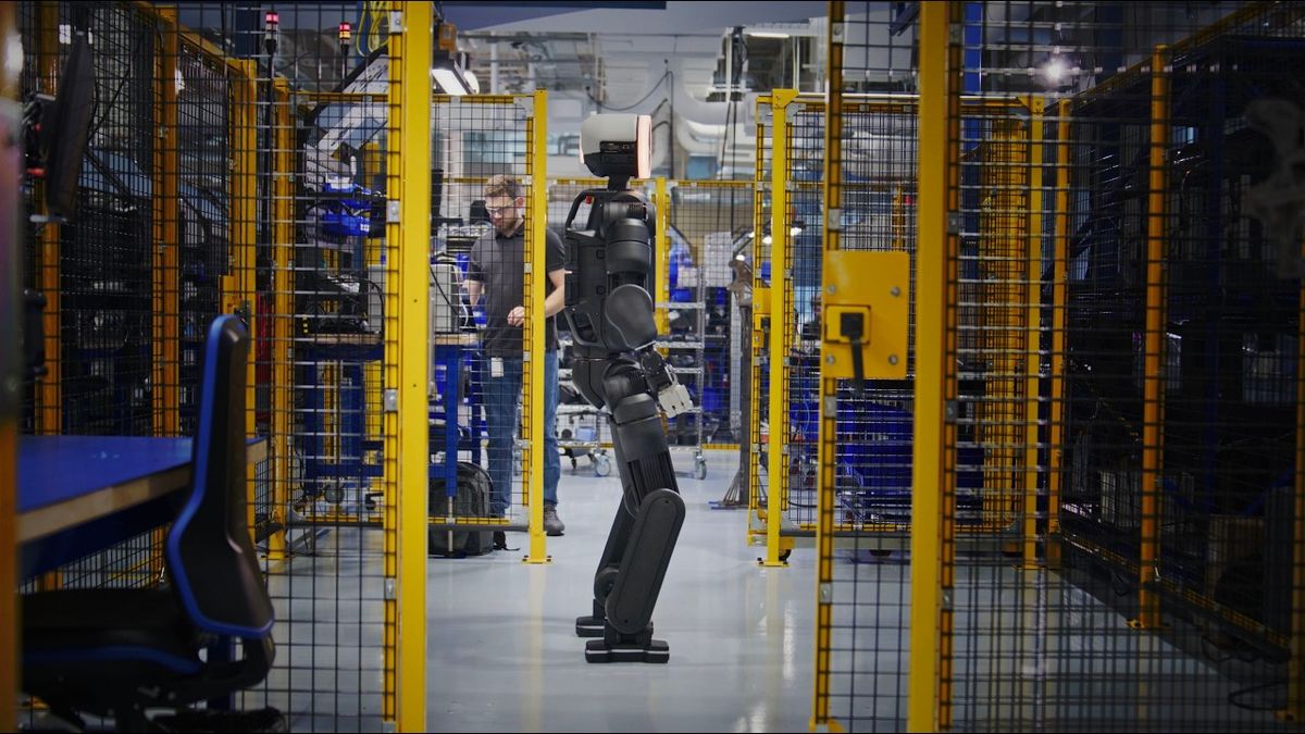 Schwarzer humanoider Roboter Atlas geht durch eine Werkhalle mit gelben Gitterzäunen, dahinter arbeitet ein Mitarbeiter an einer Werkbank