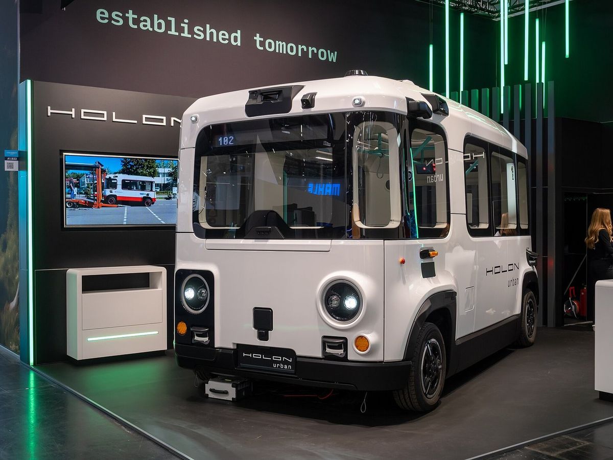 Weißer, kastenförmiger Elektrokleinbus mit runden Scheinwerfern, großer Frontscheibe und Aufschrift „Holon urban“ auf einem Messestand, darüber der Schriftzug „established tomorrow“