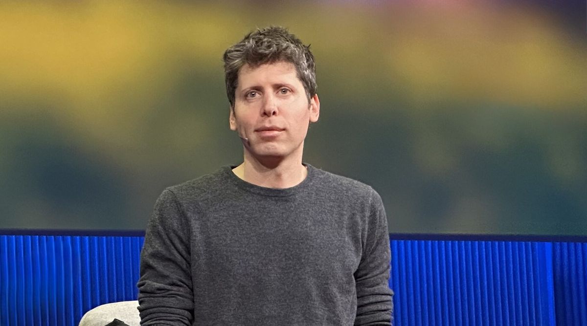 Sam Altman in dunkelgrauem Pullover sitzt mit Headset-Mikrofon auf einer Bühne, hinter ihm eine blaue Lamellenwand und eine unscharfe gelb-grüne Projektion