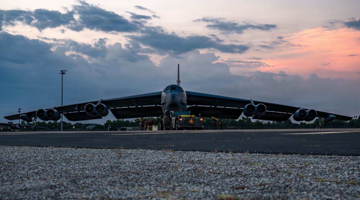 Ein achtstrahliger B-52H-Bomber steht in der Abenddämmerung frontal auf dem Vorfeld, unter dem Rumpf ein gelbes Schleppfahrzeug und mehrere Soldaten, dahinter Bäume und ein Flutlichtmast, am Himmel blaugraue Wolken mit rosa Abendrot