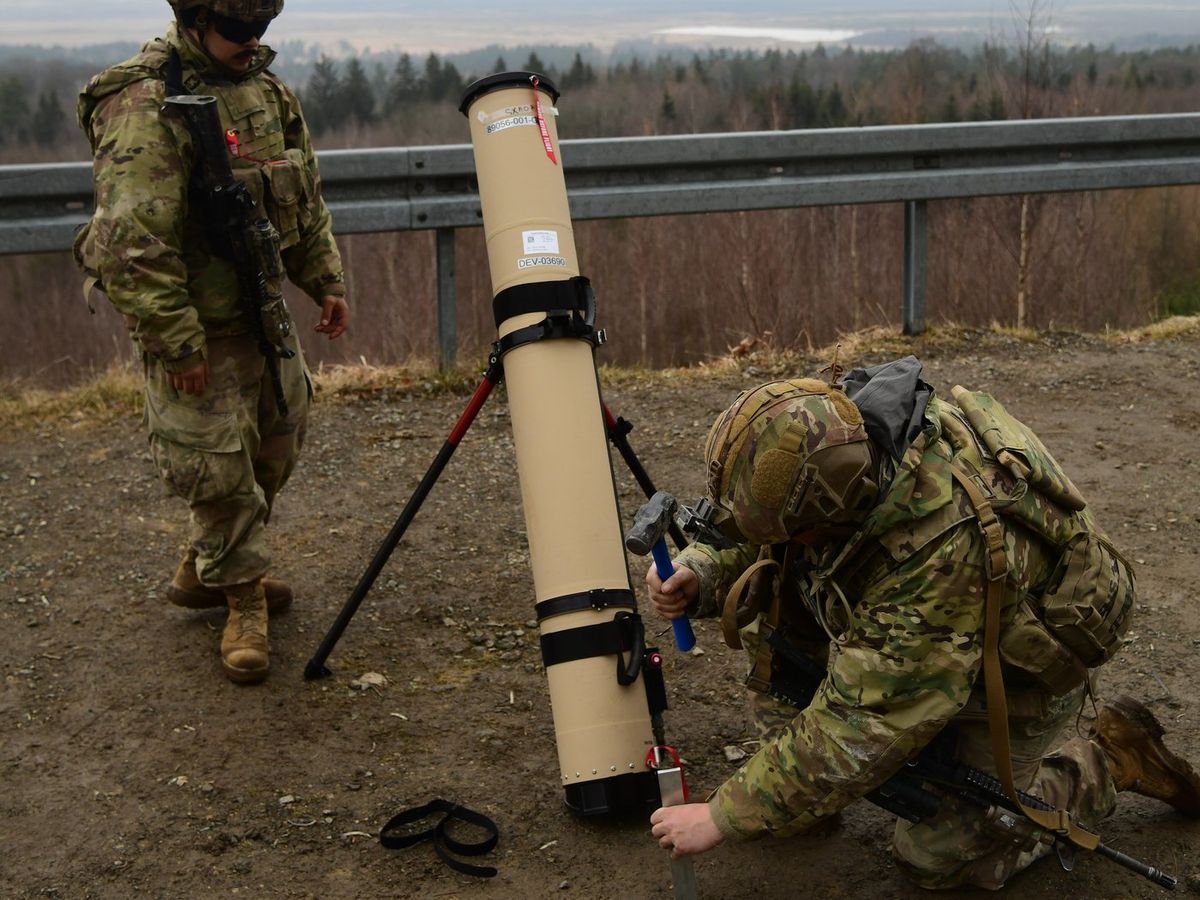 Zwei Soldaten in Tarnuniform mit Helmen an einem beigefarbenen Startrohr auf einem Dreibein, einer kniet und schlägt mit einem Hammer einen Erdanker ein, dahinter Leitplanke, Wald und eine weite Ebene