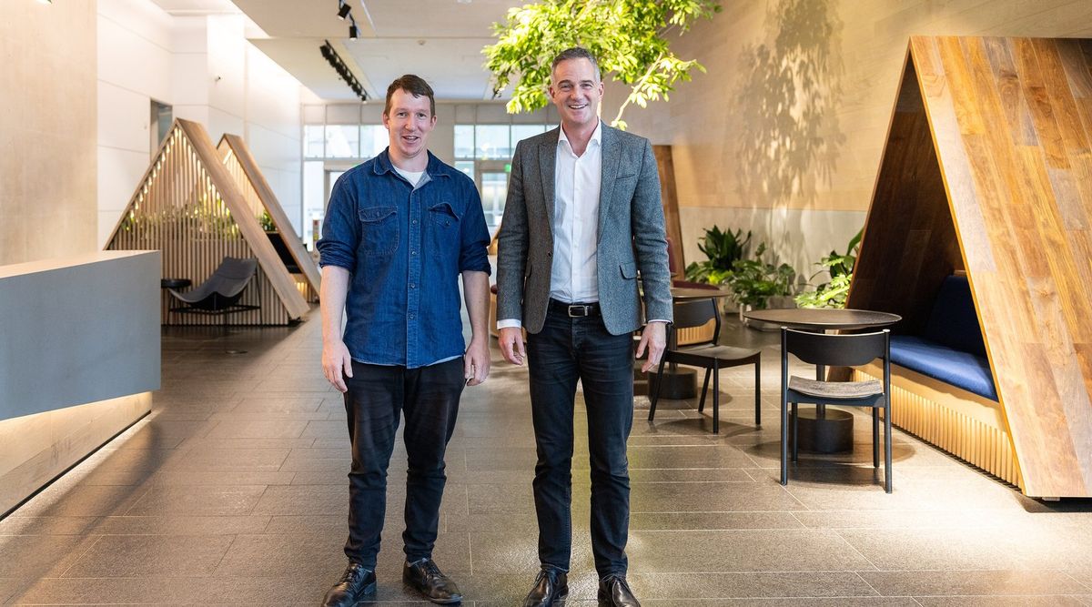 Zwei Männer stehen in einer Bürolobby mit hölzernen, zeltförmigen Sitznischen und einem Baum unter der Decke; links ein jüngerer Mann im blauen Jeanshemd, rechts ein Mann im grauen Sakko mit weißem Hemd
