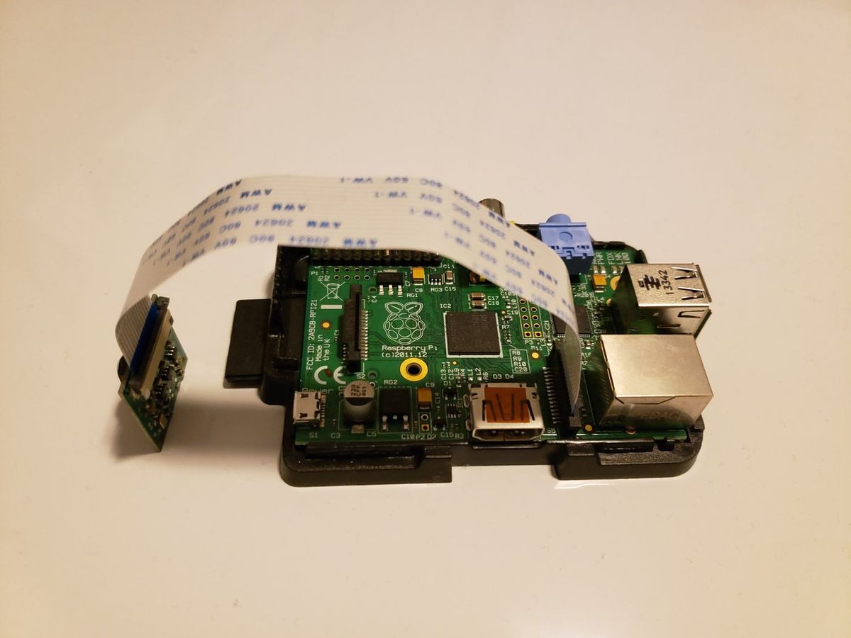 Grüne Raspberry-Pi-Platine der ersten Generation in einem schwarzen Gehäuseunterteil, daran über ein breites weißes Flachbandkabel ein kleines Kameramodul, das links aufrecht steht
